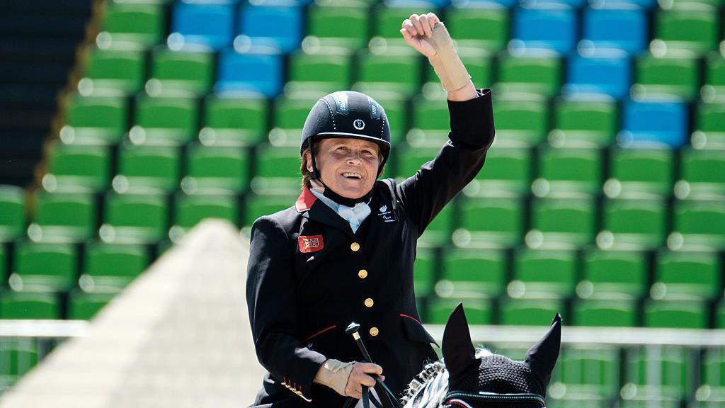 Anne Dunham is sitting on a horse with a fist in the air in black riding gear. She is smiling.