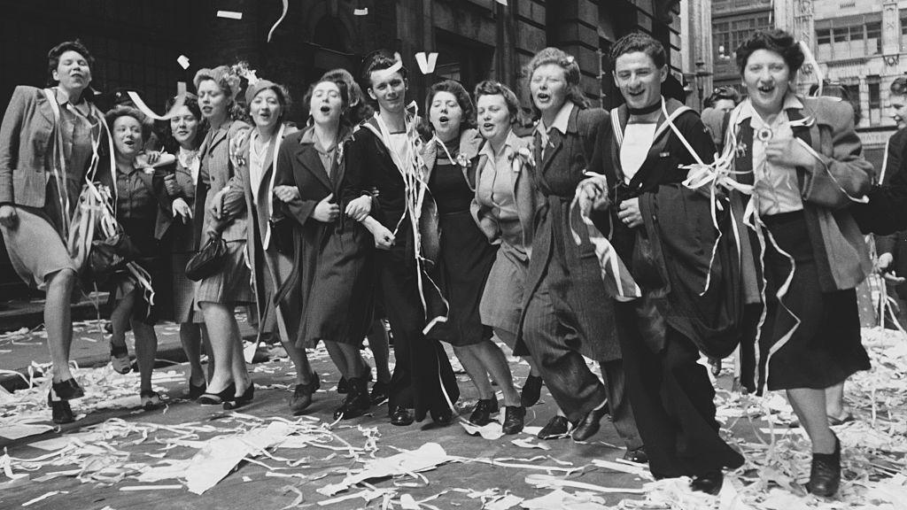 A black and white photo of a line of people linking arms, dancing and singing. They are wearing 1940s clothes and the two men are wearing sailors' uniforms. There are decorative streams of paper all over them and the street. They are in London.