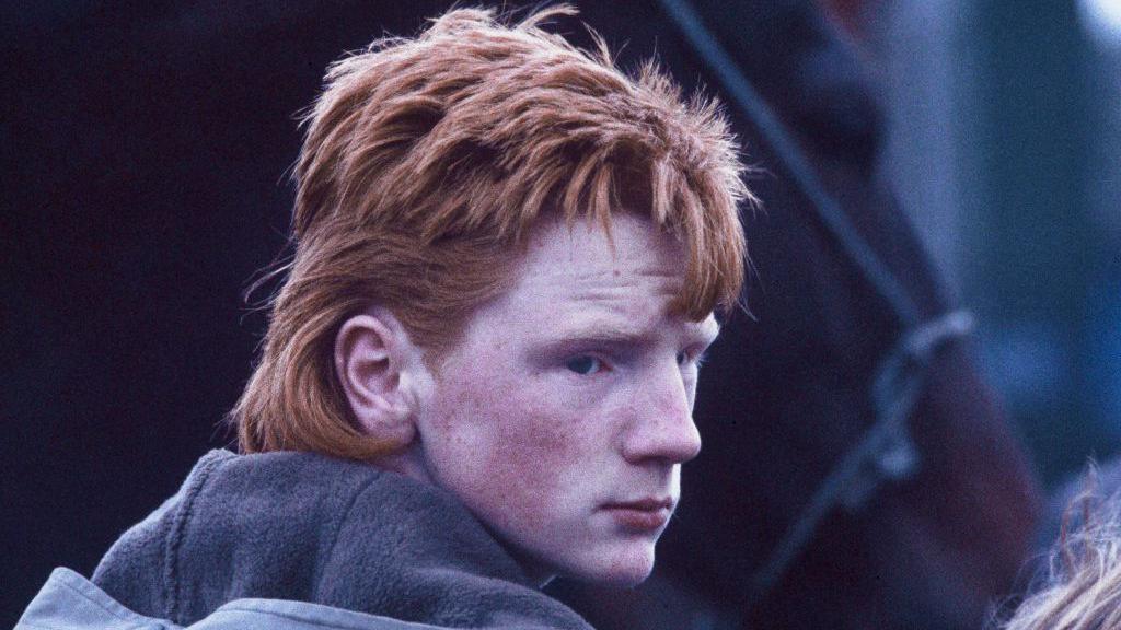 Young Irish traveller with ginger mullet, in grey hooded jacket stood in front of a brown horse