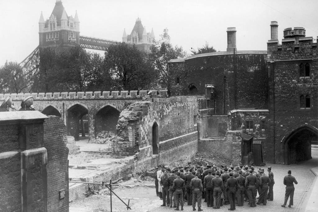 A black-and-white photo showing a group of soldiers inspecting damage to a wall in the Tower of London. One of the walls has been destroyed. Tower Bridge is visible in the background.
