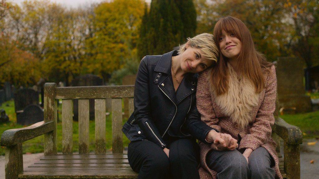 Jodie Whittaker and Aimee Lou Wood sit together on a bench, holding hands and smiling.