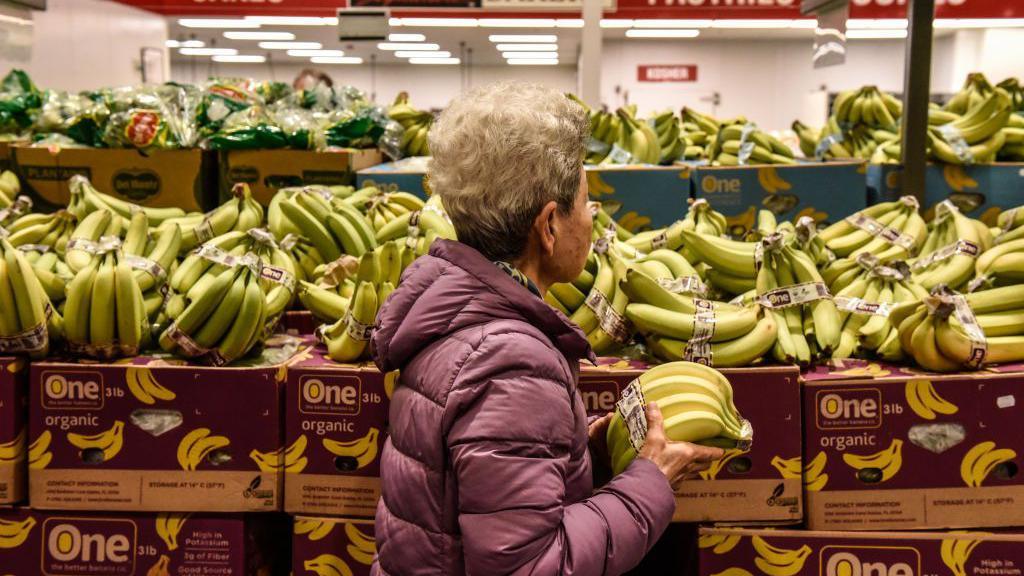 A woman holds bananas while walking through a grocery store aisle.