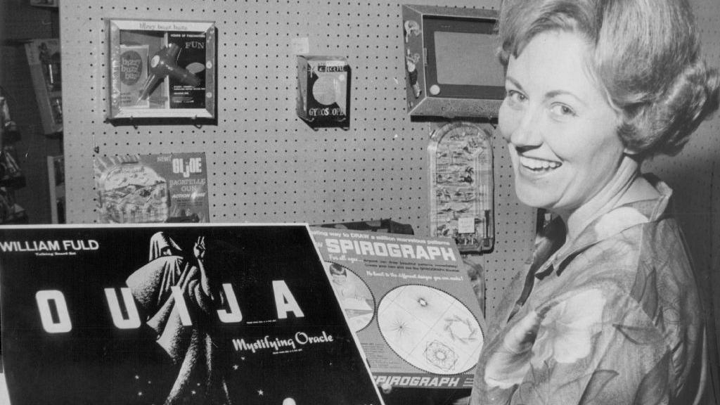 A woman with a lrage 1960s hairstyle smailes for the camera while holding a ouija board game, which had a cloaked and sinister figure on the box, and a boxed Spirograph in her other hand in this black and white image.