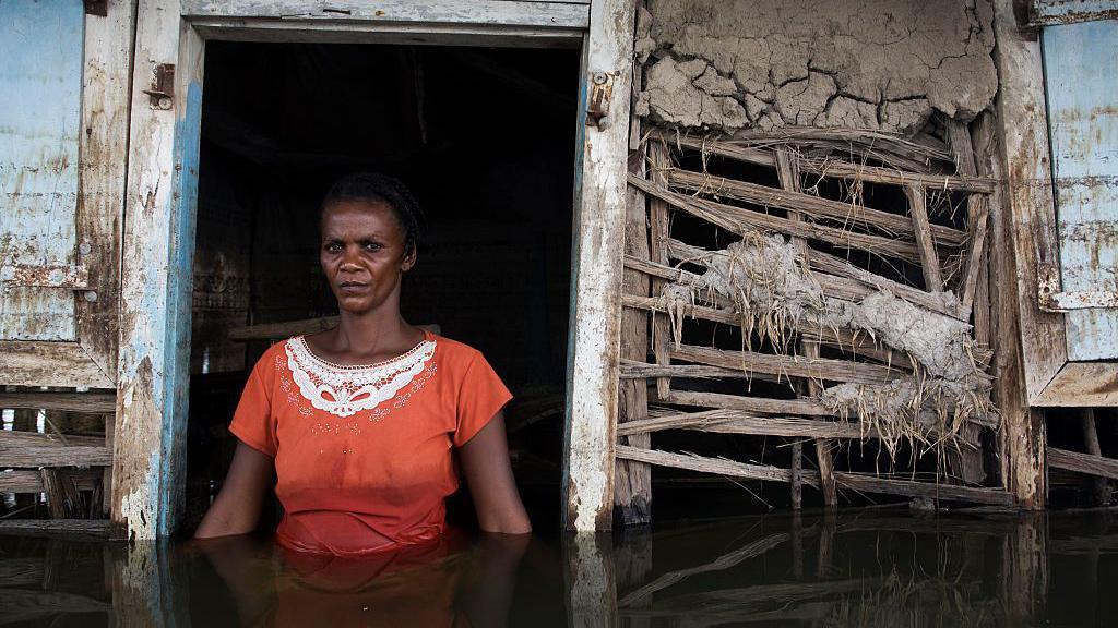 A woman in an orange dress stands in the doorway of her flooded house on the outskirts of the city of Gonaives in Haiti. The water level is at waist height. The walls of the house are substantially damaged.