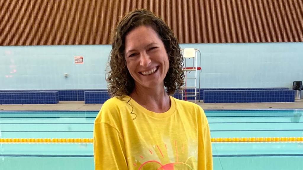 A woman with brown curly hair and a yellow tshirt stood in front of a swimming pool