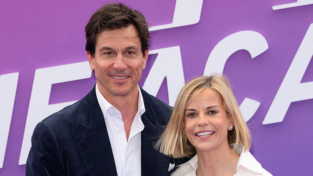 A man with shortish dark hair is wearing a black jacket and white shirt as he smiles for the cameral while standing next to a smiling woman with short blonde hair who is wearing a cream-coloured oven-neck blouse