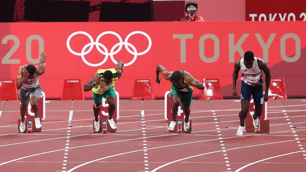 Reece Prescod is disqualified after a false start during the men's 100m semi-final. He has risen from his blocks on the track and is wearing the Team GB kit - a white vest with navy trunks. Three other runners are coming out of their blocks in the lanes alongside him. The Olympic logo can be seen on a red board behind them along with the words "Tokyo 2020".