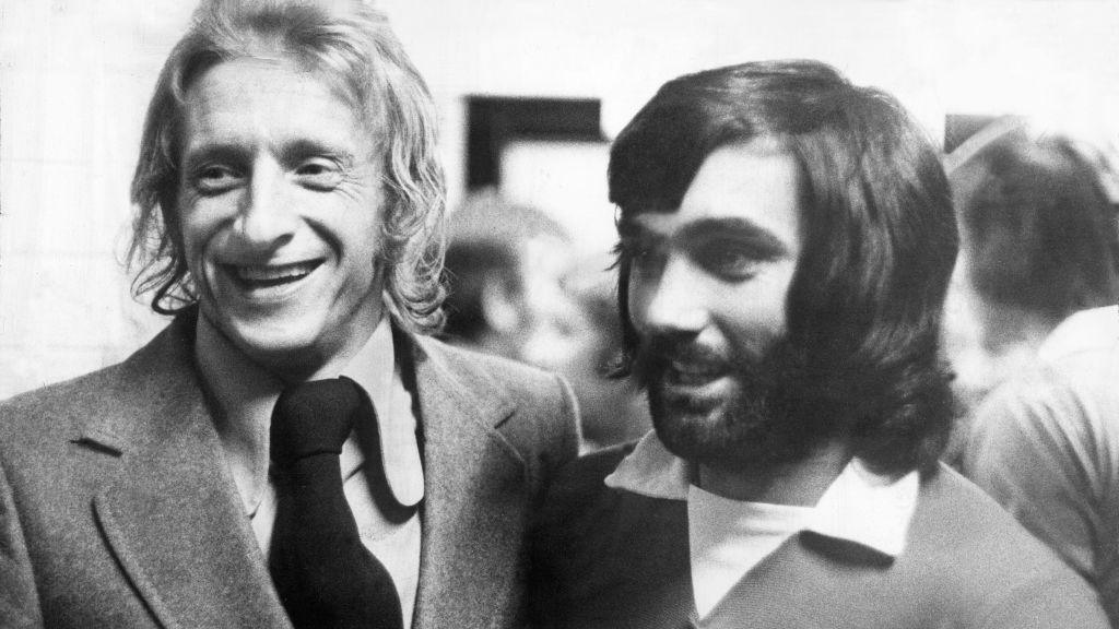 A black and white picture of Denis Law in a suit with long fair hair and George Best with long dark hair and stubble wearing a shirt and jumper.