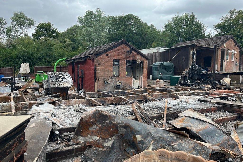A burnt-out outhouse building, made of brick, is in the background, and behind it are trees. In the foreground is the devastation of fire damage - charred debris.