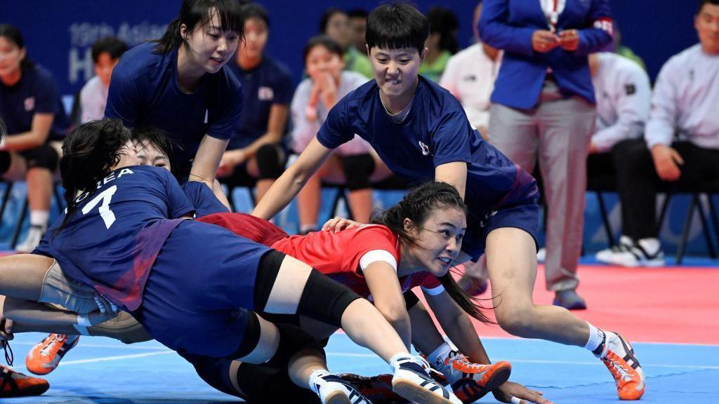 One Kabaddi player is diving to the floor in a red t-shirt. Around her, trying to tackle her are four Kabaddi players in navy blue.