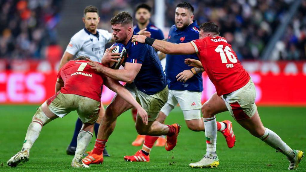 A rugby player dressed in a blue shirt being tackled on a rugby pitch by two players dressed in red shirts