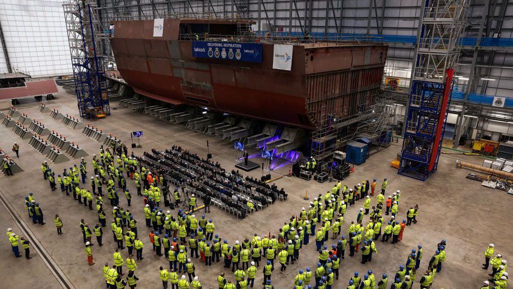 Crowds of people in fluorescent yellow safety jackets and hard hats viewed from above. They are in a large warehouse looking at a section of a brown warship in construction.