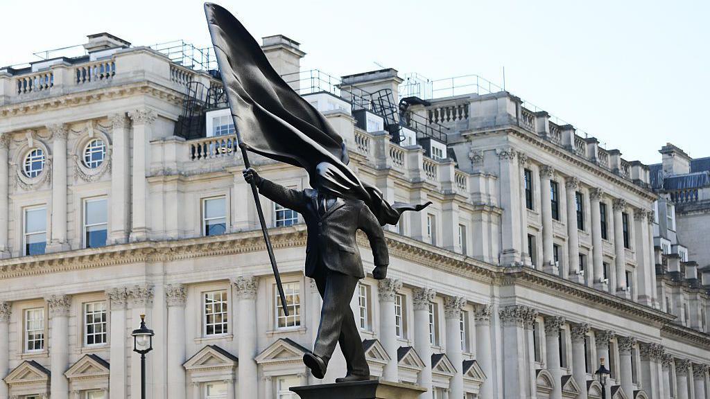 the statue of a man in a suit walking off a plinth holding a flag that is covering his face.