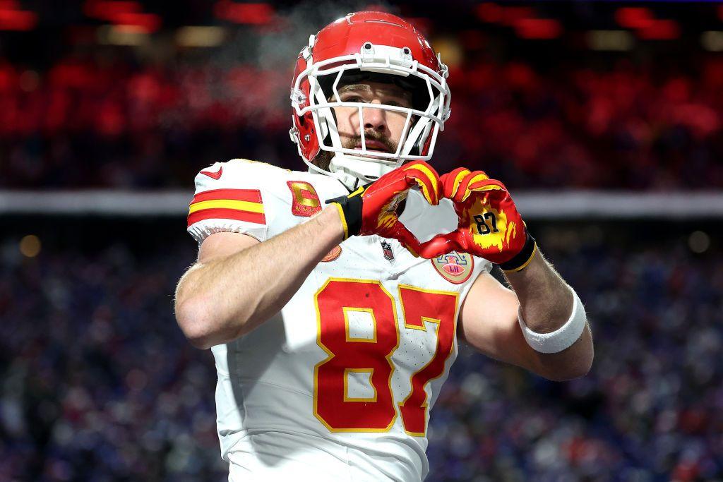 Travis Kelce in his number 87 Kansas City Chiefs shirt. He celebrates making a heart shape with his hands.