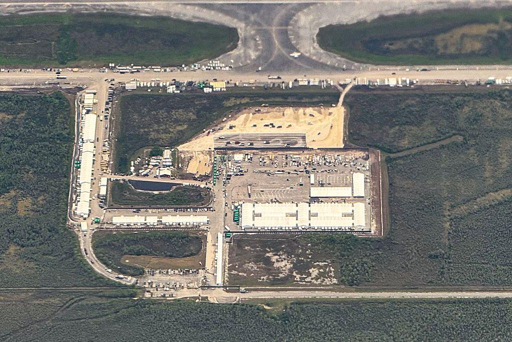 An aerial view of a migrant detention center, dubbed "Alligator Alcatraz," showing large areas of green and swamps with patches of tarmac and concrete airfield in the middle with buildings and vehicles seen on it.