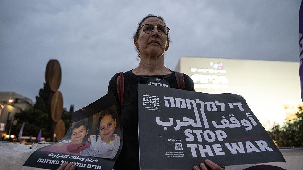 A woman stands in protest holding up two signs - one showing the image of two Gazan children killed in the war and another that says in Hebrew, Arabic and English: "Stop the War"