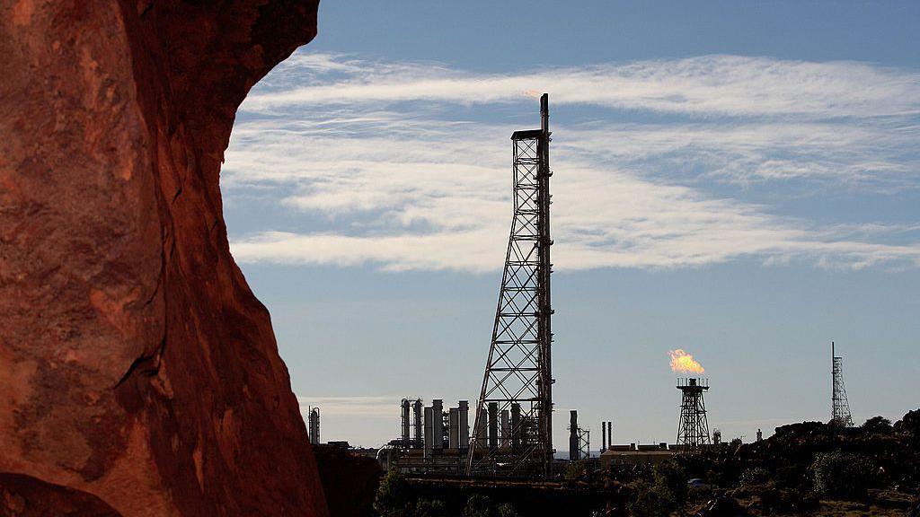 A red rock shelf in the foreground with an industrial site in the background, including a flaming gas tower