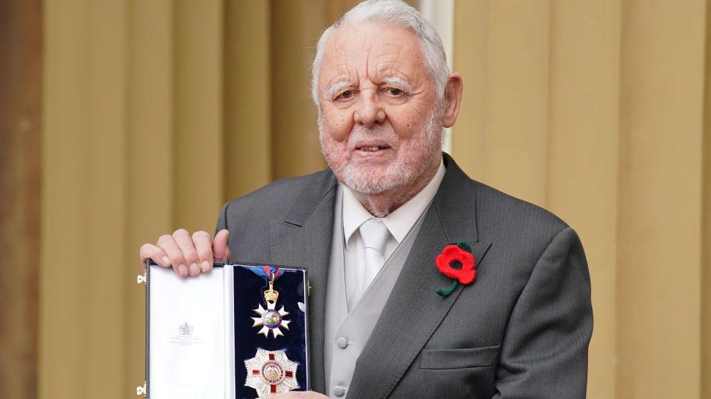 Sir Terry pictured outside Buckingham Palace holding his Knighthood in his hands toward the camera. He is smiling. He has grey hair and a grey beard. He is wearing a dark grey suit with a lighter grey waistc oast and a white shirt and white tie. He has a red poppy in his left hand breast for Remembrance Day. 