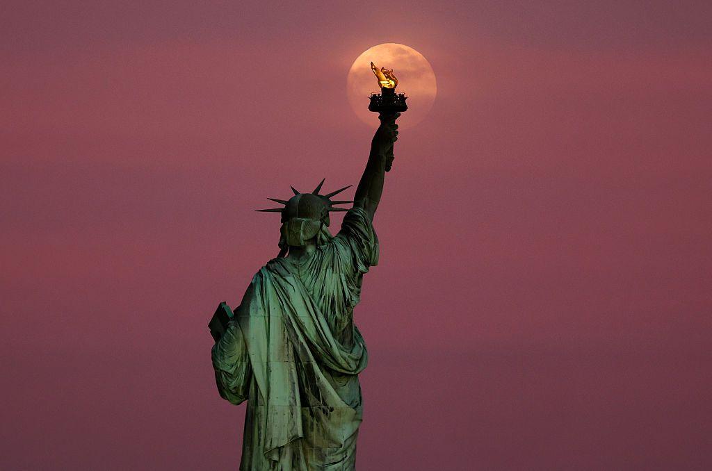 The Flower Moon rises through clouds behind the Statue of Liberty as the sun sets in New York City on May 11, 2025, as seen from Jersey City, New Jersey.