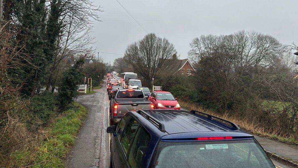 The same traffic jam is pictured from the opposite side of the road. The road has grass verges and narrow pavements on either side. 