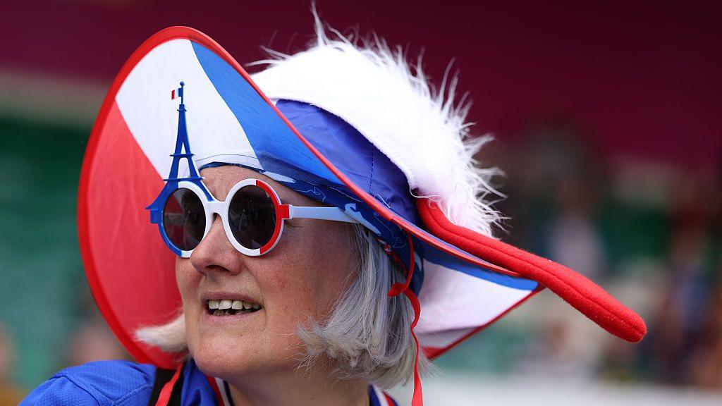 A France fan wearing a red white and blue hat and red white and blue sunglasses with an Eiffel tower on one lens. She is looking to the side.