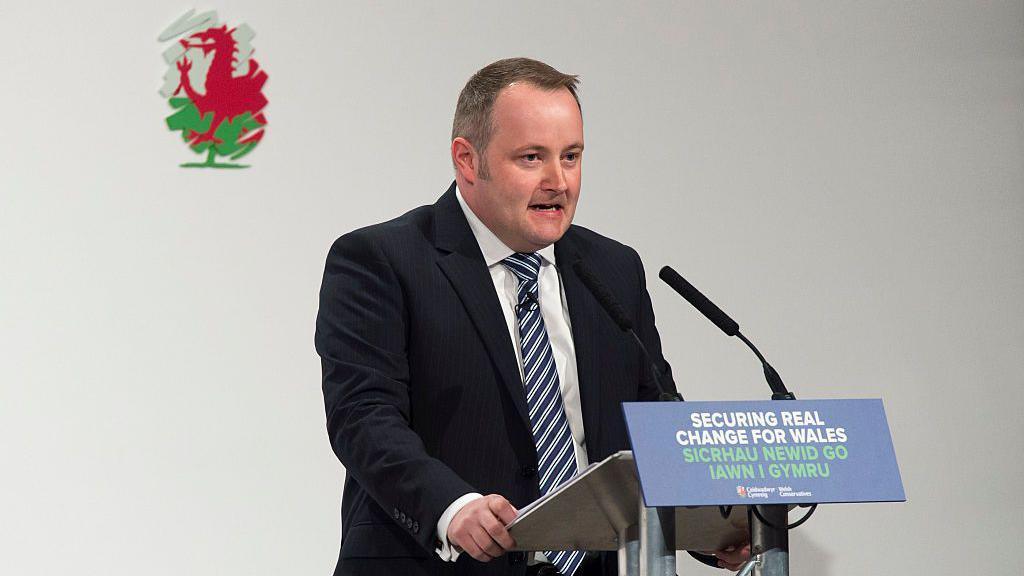 A man is stood in front of a lectern. He is wearing a blue suit with a white shirt underneath and a blue stripy tie