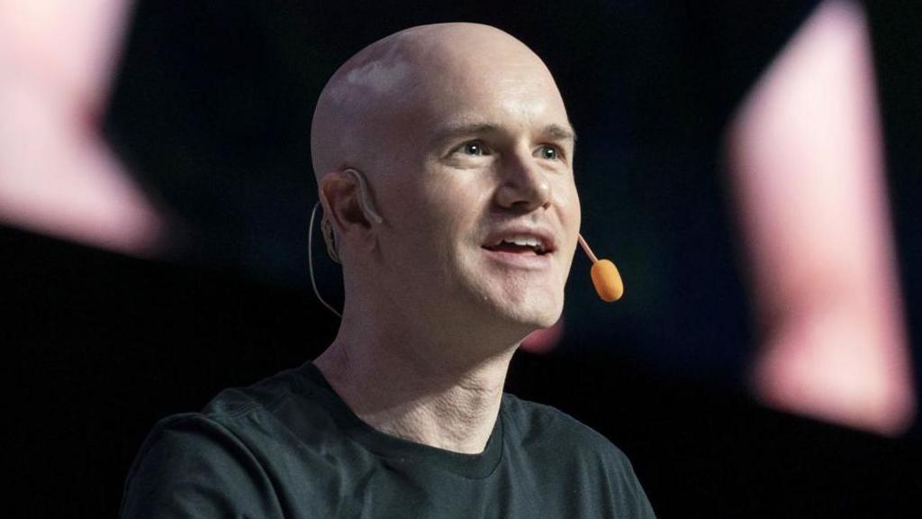 Coinbase chief executive Brian Armstrong shown speaking on stage at an event, wearing a black t-shirt and a microphone.