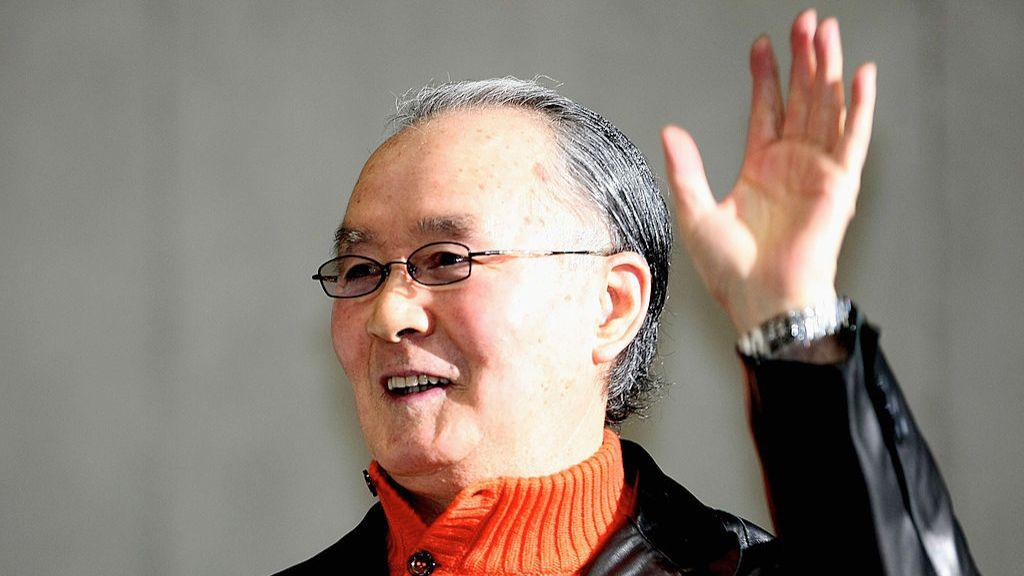 A photo of Shigeo Nagashima wearing a orange turtle neck jumper and a black leather jacket. He is waving.