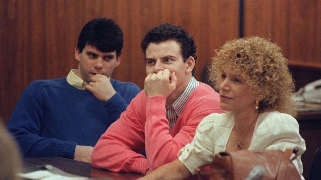 Erik and Lyle Menendez sit with their attorney in a courtroom in Los Angeles County. They are each wearing colourful sweaters 