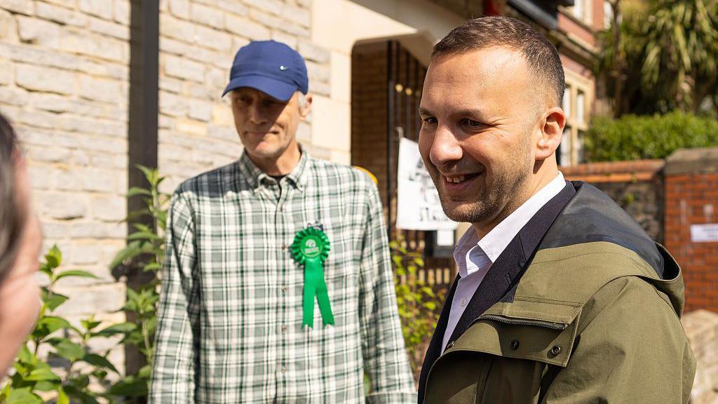 Zack Polanski in a green coat next to man wearing a welsh Green Party rosette