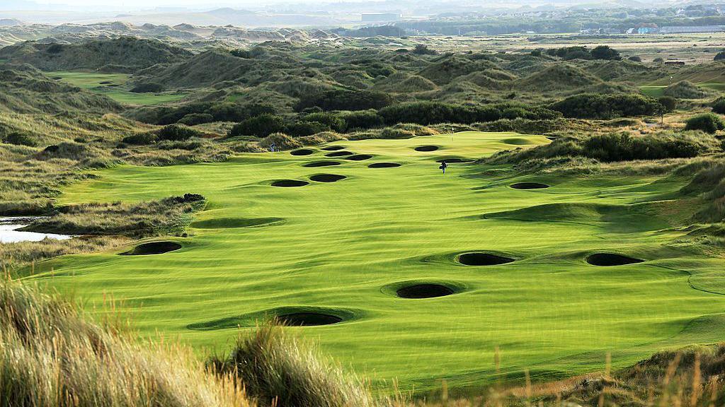 The 18th hole at Trump International Golf Links at Balmedie in Aberdeenshire. We see a verdant, undulating fairway pocketed by deep bunkers. The hole lies among large, deep grassed-over dunes. A lone golfer can just be seen in the far distance, approaching the green.