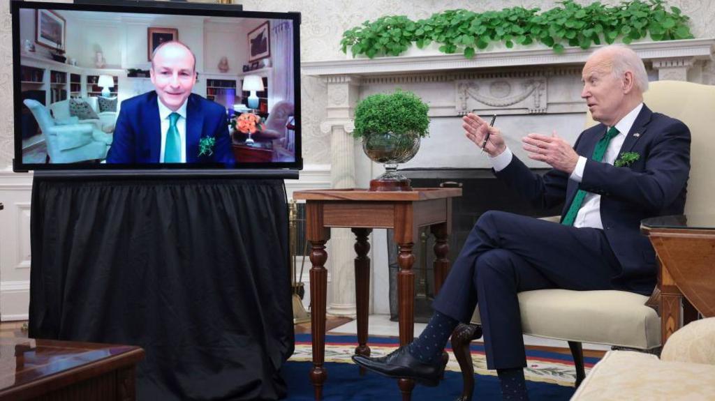 Micheál Martin is seen on a TV screen in the Oval Office in a blue suit and green tie, next to the TV is a bowl of shamrock on a mahogany table and next to that is Joe Biden in a dark suit, white shirt and green tie.