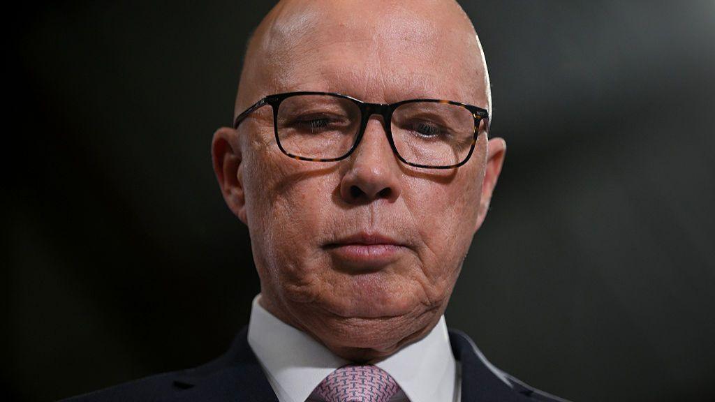 Peter Dutton, wearing glasses and a suit, looks down, his face neutral, in a close-up headshot