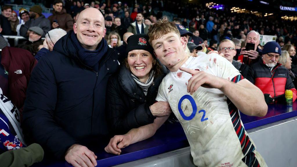 Andrew Smith with very short hair, smiling and wearing a dark anorak, stands next to Judith, who is wearing a blue hat with an England rose logo. They are both behind the crowd barrier at a stadium. Fin, with short ginger hair, wearing a white England strip with an O2 logo, is standing on the other side of the barrier with his arm reaching towards Andrew's hand.