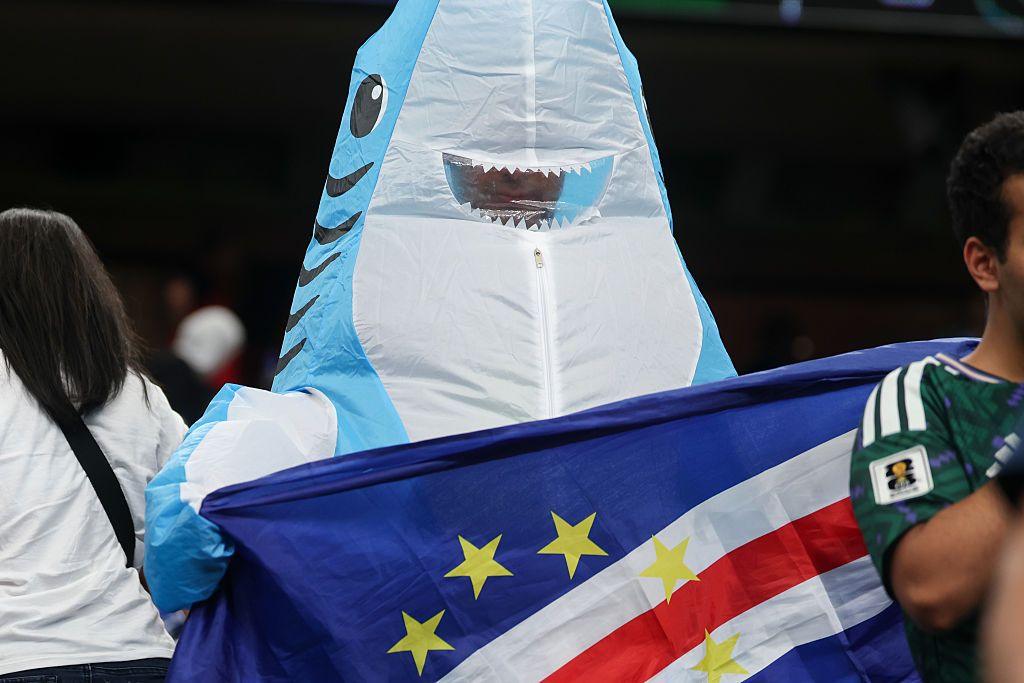 A Cabo Verde fan wears a blow up shark costume and holds the Cabo Verde flag .