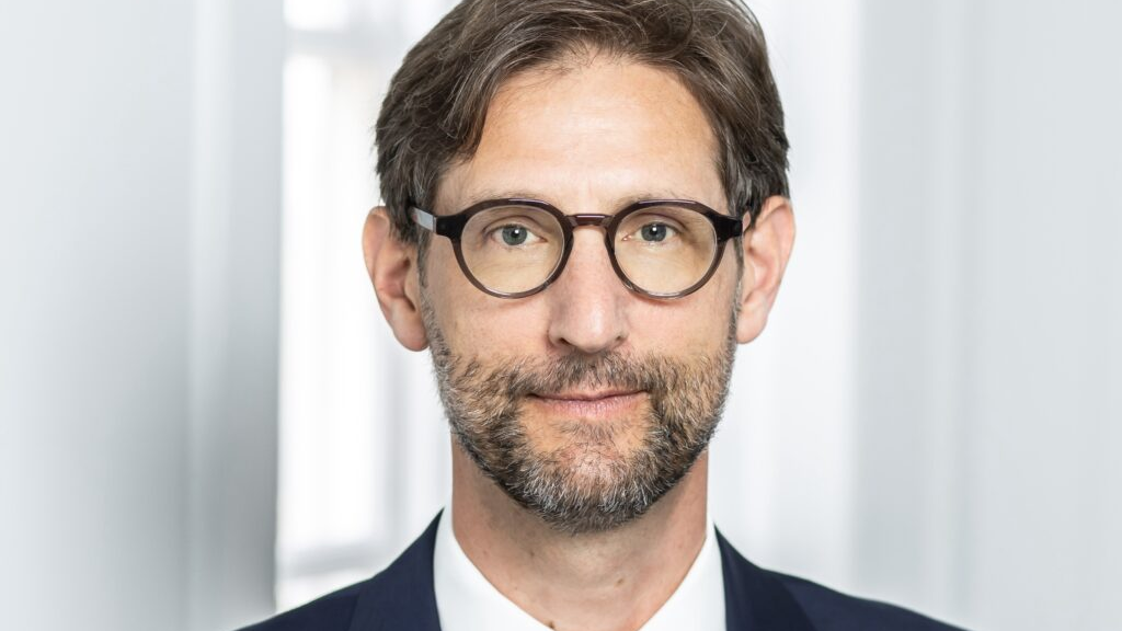 Nicholas Griffin KC, wearing a dark blue suit and white shirt. He has round dark-rimmed spectacles and short beard. He has short dark hair and is stood in front of a light blurred background. He is looking directly at the camera.