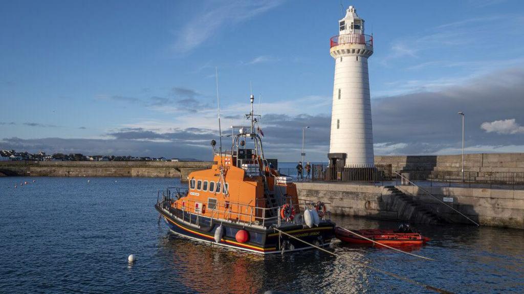 Donaghadee Harbour with a white lighthouse at the end of one half of the harbour walls. In front is an orange and navy blue lifeboat. The harbour waters are calm and dark blue. Beyond the harbour walls is the sea and an expanse of low stratus clouds. It's feels like a sunny evening.