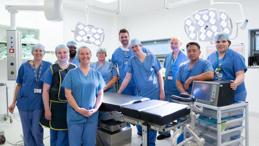 10 medics, all of them wearing blue gowns pose next to a bed in an operating theatre.