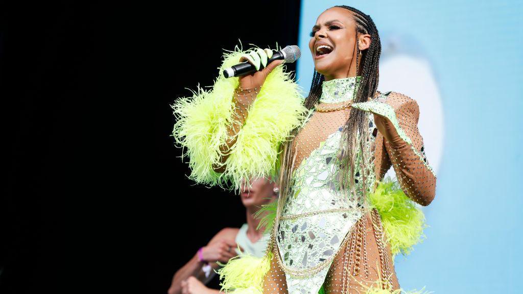 Samantha Mumba with long, braided hair, wearing a green bodysuit with chain and feather details. She is singing into a microphone.