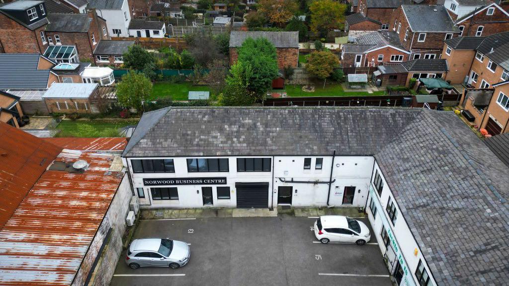 An aerial view of the Hart Space studio, located in a two story building with white painted walls and black-framed windows. The building forms an L-shape around a courtyard used as car parking. On the left-hand side of the image, the courtyard is enclosed by a building with a rusty corrugated roof, where the garage next door was located.