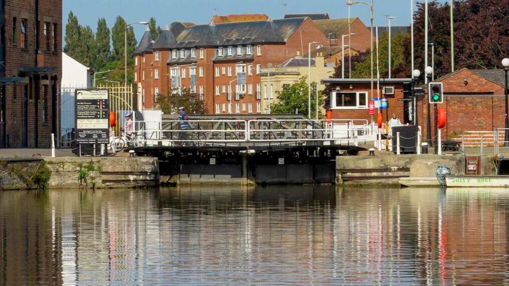 Gloucester Lock. Gloucester Lock. To the left is a white building and behind that a brick building. On the right is a low brick building. In the foreground is water