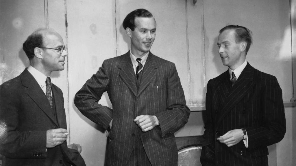 Black and white image James Sayers (centre) with Professor John Randall (left) and Professor Henry Albert Howard Boot. John Randall is wearing glasses and a dark suit and holding a cigarette. James Sayers is wearing a grey pin-stripe suit. Henry Albert Howard Boot is wearing a dark suit. All three are engaged in conversation.