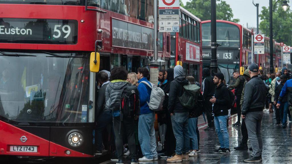 A queue of people stand in a rainy London street waiting to board a bus. There are bus stops and other buses visible in the background.