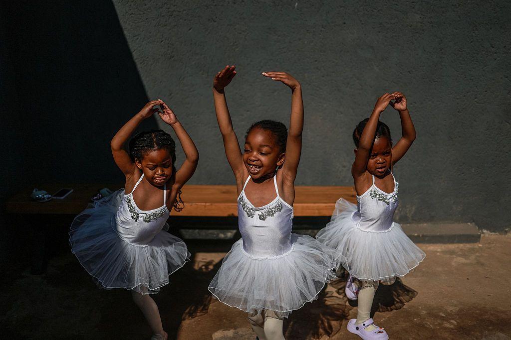 Three girls wearing white leotards and tutus stretch their arms out.
