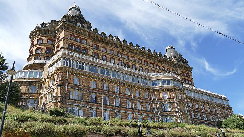 The exterior of a large, Victorian-era hotel, viewed from below. The brickwork is light-coloured, with white windows and a wraparound balcony with white railing around the mid-section of the building.