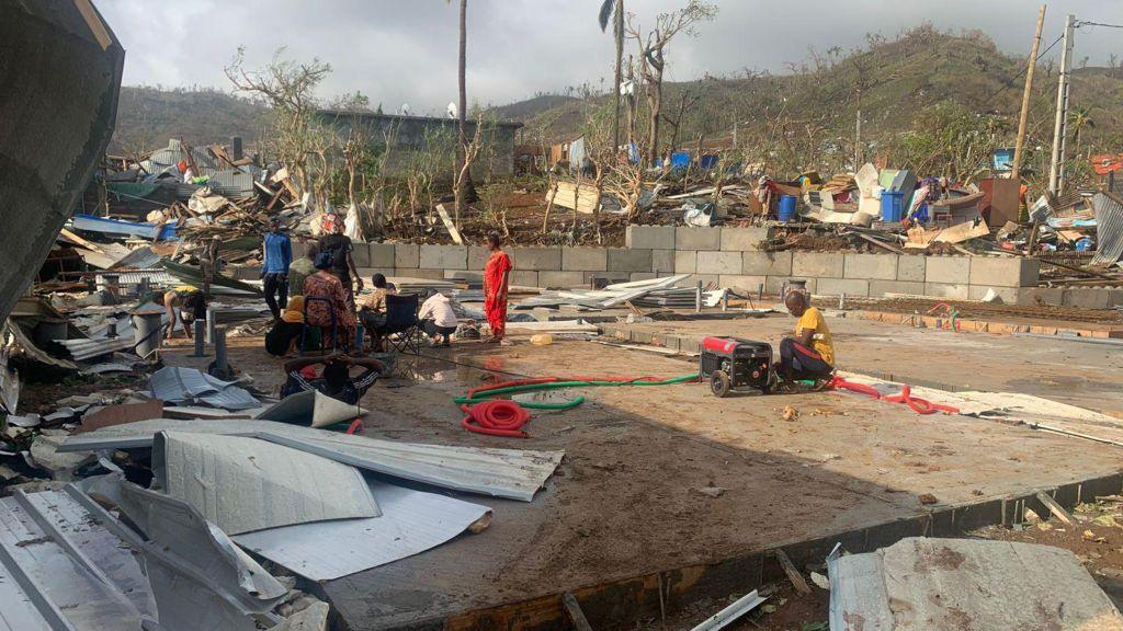 People stand or crouch on the ground beside debris and destroyed homes in Mayotte