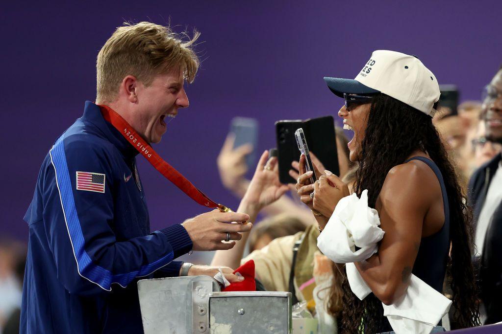 hunter woodhall and tara davis-woodhall beam at one another across a barrier as hunter holds up his gold medal to show tara and tara holds up her phone in the crowd