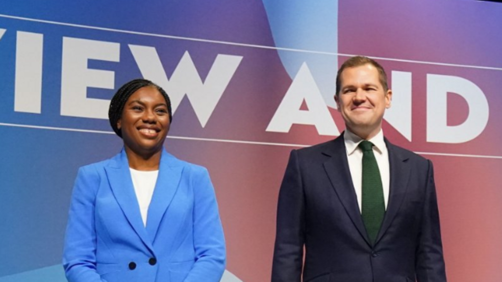 Kemi Badenoch in bright blue suit and white shirt stands next to Robert Jenrick in dark-blue suit, white shirt and forest-green tie