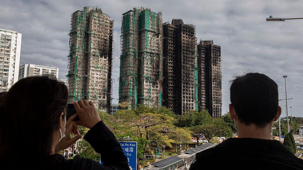 The back of two people's heads as they look at four charred high-rise blocks in the distance, with trees and roads between them and the towers
