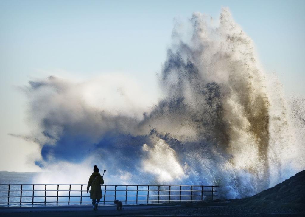 A walker and dog are silhouetted in black against a huge wave striking the sea front at Whitley Bay. The wave is coloured blue at the bottom and white higher up